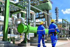 Dos trabajadores con cascos y uniformes azules de seguridad inspeccionan equipo en una plataforma elevada dentro de una planta industrial. El fondo muestra una extensa red de tuberías, tanques y estructuras metálicas bajo un cielo azul con nubes blancas.