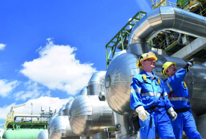 Dos trabajadores con cascos amarillos y uniformes azules de seguridad inspeccionan equipo en una plataforma elevada dentro de una planta industrial. El fondo muestra una extensa red de tuberías y tanques metálicos brillantes bajo un cielo azul con nubes blancas dispersas.
