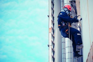 Un trabajador vestido con un traje azul y casco rojo escala una estructura alta usando cuerdas y arnés de seguridad, con un cielo claro de fondo.