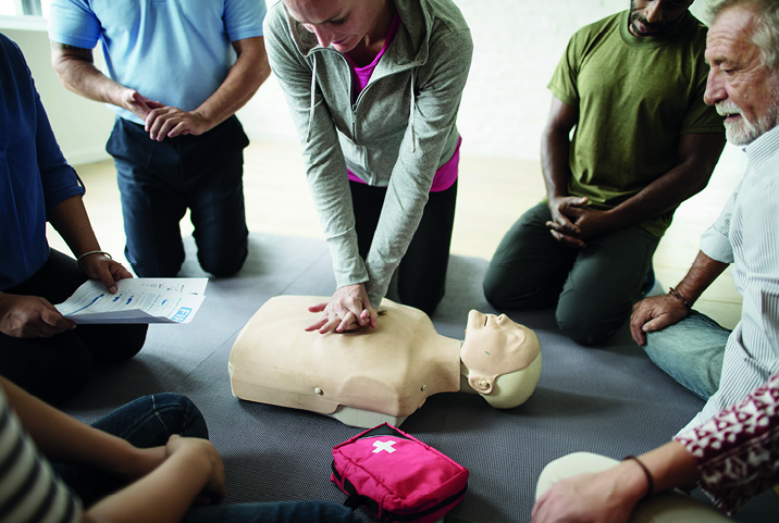 Un grupo de personas sentadas en círculo realiza un entrenamiento de RCP en un maniquí, con una instructora practicando compresiones torácicas y un kit de primeros auxilios visible.