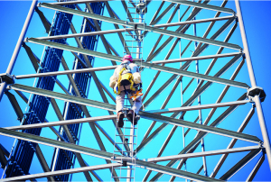 Un trabajador con casco y arnés asciende una alta torre de comunicación de acero bajo un cielo azul, utilizando una escalera integrada en la estructura.