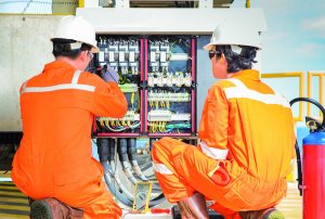 Dos técnicos con trajes naranjas y cascos blancos reparan un cuadro eléctrico con cables y equipos electrónicos, en un entorno industrial con un extintor cercano.