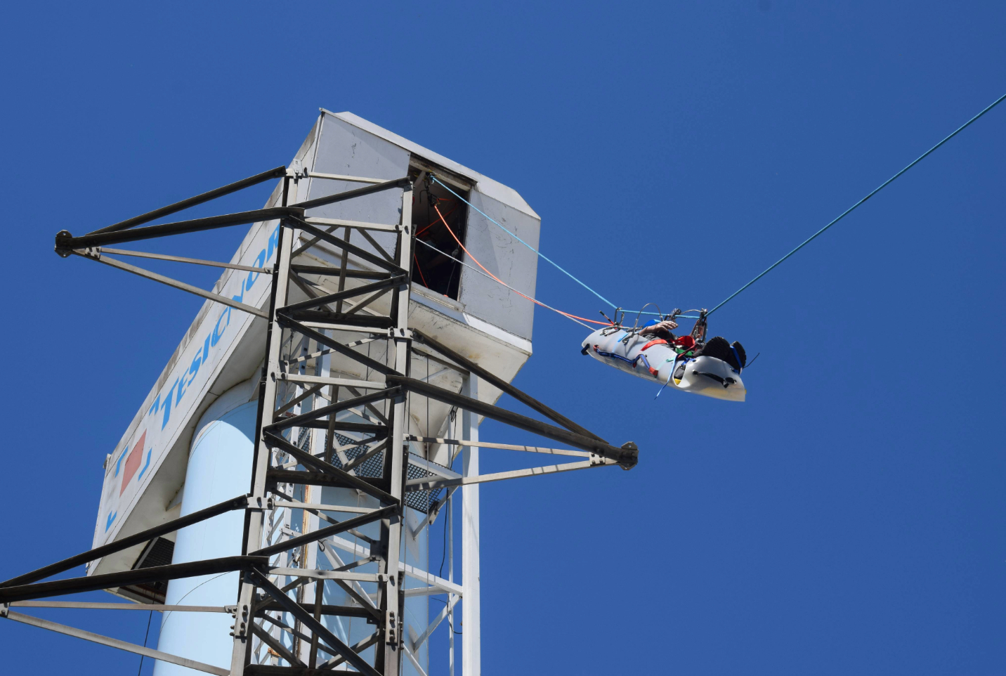 Una persona con arnés siendo lanzada desde una torre alta con el logotipo de "Tesiscnor", bajo un cielo azul despejado, durante un curso de formación GWO ART.