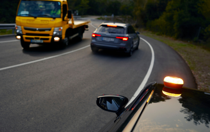 Vehículo detenido con luz ámbar de emergencia en el techo en una curva de carretera, con un coche y una grúa acercándose al fondo.